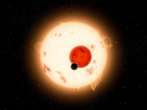 Artistic view of the components of the ternary system Kepler-16 in transit. The late-K star (large and orange) and M-dwarf star (reddish and smaller) and the exoplanet (dark) are shown to their relative size at the time of a transit. Sunspot activity as seen on Kepler-16A was indeed detected by Kepler.